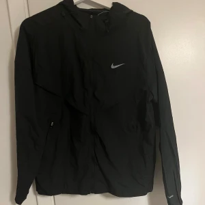 Svart Nike vindjacka med huva - Svart vindjacka från Nike med huva och dragkedja framtill. Jackan har ett stilrent Nike Swoosh-tryck på bröstet, två sidofickor med dragkedja och justerbar dragsko vid huvan. Perfekt för dig som gillar sportig och clean stil.