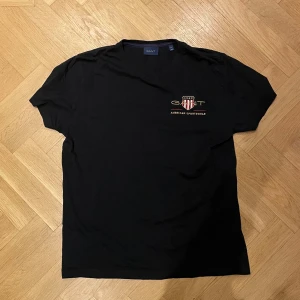 Svart GANT t-shirt med broderad logga - Svart t-shirt från GANT med broderad logga och sköld på bröstet. Klassisk passform och rund halsringning. Snygg detalj med texten 'American Sportswear' under loggan. Perfekt för en clean och sportig look. Använd sparsamt och därav i väldigt bra skick. 