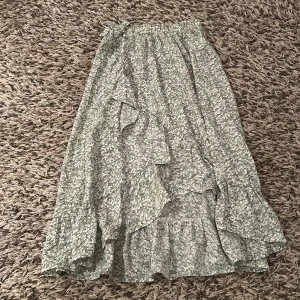 Blommig långkjol med volang  - Supersöt långkjol i ljusgrönt med vitt och grönt blommönster. Kjolen har volangdetaljer och en slit längst ner. Perfekt för dig som gillar en stilren look men med något extra💗