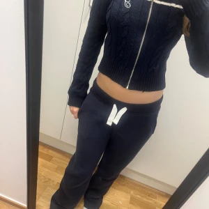 Mörkblå stickad kofta från STR Teen - Snygg mörkblå stickad kofta från STR Teen med vita detaljer vid krage och bröst, samt broderad bokstav. Croppad modell med kabelstickat mönster, dragkedja framtill och långärmad. Perfekt för dig som gillar preppy stil och vill ha något som sticker ut.