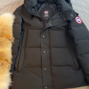 Svart dunjacka från Canada Goose M - Svart dunjacka från Canada Goose i storlek M. Jackan har en klassisk puffer-design med huva och avtagbar pälsdetalj. Ikonisk röd och vit logotyp på ärmen, dragkedja och tryckknappar framtill. Perfekt för kalla vinterdagar och riktigt snygg streetstil. Storlek M. Har haft den bara förra vintern några gånger då ja har också en Burberry jacka så jag brukar ha ibland denna o andra jackan alltså förra året. Inget fel på den- ingahål eller fläckar helt ny silren med scan o allt, pälsen följer med.