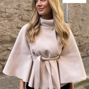 Beige kappa med bälte och capeärm - Säljer en beige kappa med capeformade ärmar och högt krage. Jackan har ett snyggt bälte i midjan och en dekorativ knapp vid halsen. Perfekt för dig som gillar stilrena och eleganta plagg med lite extra edge.