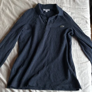 Mörkblå pikétröja Lacoste slim fit - Snygg mörkblå långärmad pikétröja från Lacoste i slim fit-modell. Klassisk krage med knappar och den ikoniska krokodilloggan broderad på bröstet. Perfekt för dig som gillar stilrena och sportiga plagg med skön passform.