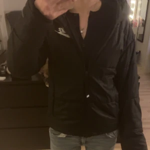 Svart pufferjacka från Nike - Svart pufferjacka från Nike med huva och vit logga på bröstet. Jackan har dragkedja framtill och är quiltad för extra värme. Perfekt för kalla dagar och har en clean, sportig look. Modellen är långärmad och har en normal passform.