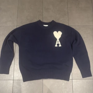 Mörkblå stickad tröja från Ami Paris - Snygg mörkblå stickad tröja från Ami Paris med ett vitt hjärta och bokstaven A broderat på bröstet. Tröjan har rund hals, ribbade muddar och lång ärm. Perfekt för dig som gillar stilrena detaljer och vill ha något som sticker ut lite extra.