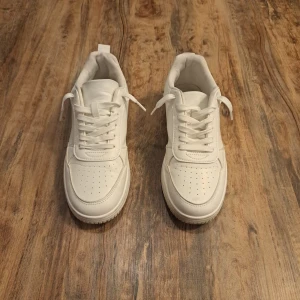 Vita Sneakers  - Stilrena helvita sneakers från F&F med klassisk design, rund tå och perforerade detaljer på tån. Skorna har platt sula, snörning och är tillverkade i syntetmaterial. Perfekta för dig som gillar en clean och fräsch look.