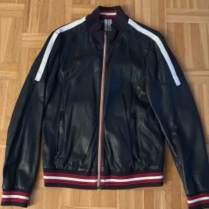 Svart skinn bomberjacka Oakwood - Snygg svart bomberjacka i skinn från Oakwood med vita ränder på axlarna och coola röda, vita och marinblå ribbade muddar. Jackan har dragkedja framtill, två fickor med dragkedja och sportiga detaljer på insidan. Perfekt för dig som vill ha en stilren och edgy look.