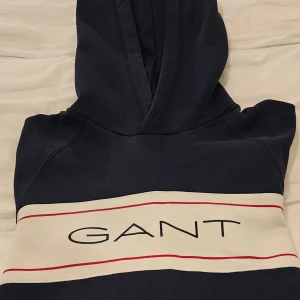 Marinblå hoodie från GANT, S - Snygg marinblå hoodie från GANT i storlek S. Tröjan har en huva och ett brett vitt band över bröstet med GANT-loggan och röda linjer. Perfekt för dig som gillar klassisk streetstyle och vill ha en bekväm och stilren look.