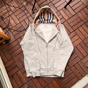 Grå hoodie med dragkedja - Snygg grå hoodie i märket Burberry i bomull med klassiskt rutigt foder i huvan. Tröjan har dragkedja framtill, snörning vid huvan och ribbade muddar. Perfekt för dig som vill ha en stilren och lyxig vardagslook. Nytt skicka använd 2 gånger.