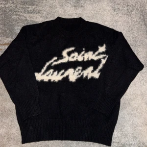 Svart stickad Saint Laurent tröja - Svart stickad tröja från Saint Laurent med vit, suddig logga framtill. Tröjan har rund halsringning, ribbade muddar och en loose fit som ger en chill vibe. Perfekt för dig som vill ha något både stilrent och edgy.