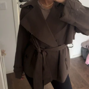 Brun trenchcoat med bälte - Snygg brun trenchcoat med bred krage och knytbälte i midjan. Jackan har dubbelknäppning framtill, långa ärmar och en klassisk trench-design som ger en cool och stilren look. Perfekt för dig som gillar tidlösa och trendiga plagg. Säljer då den är för stor för mig. Använt en gång.
