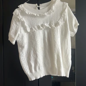 Vit blus med spetsvolang och kort ärm - Supersöt vit blus med korta ärmar, ribbad krage och spetsvolang längs halsringningen och axlarna. Blusen har hålmönster och knäppning bak i nacken. Perfekt för dig som söker en vit blus!.