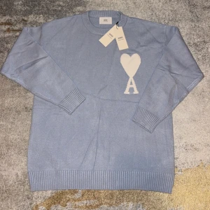 Ljusblå stickad tröja från Ami - Säljer en ljusblå stickad tröja från Ami med rund halsringning och ribbade muddar. På bröstet finns ett stort vitt hjärta med bokstaven A, vilket ger en unik och trendig look. Perfekt för dig som gillar stilrena detaljer och vill sticka ut.
