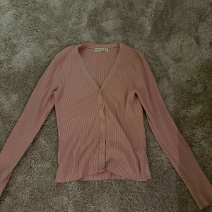 Rosa ribbad kofta  - Säljer en ljusrosa ribbad kofta från Primark i storlek XS. Koftan har v-ringning, långa ärmar och knappar framtill. Perfekt att slänga över en topp för en chill look. Materialet är mjukt och stretchigt, vilket gör den skön att bära.