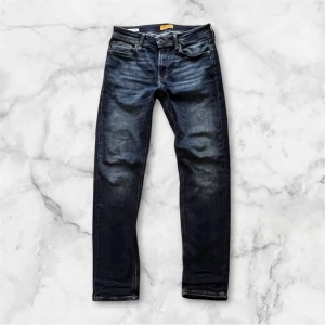 Jack & Jones mörkblå jeans Clark 30/32 - Snygga mörkblå jeans från Jack & Jones, modell Clark Regular. Klassisk femficksdesign med slitningar och kontrastsömmar. Jeansen har rak passform och normal midja, tillverkade i slitstarkt denim med läderpatch bak. Nypris 899kr: Mitt pris 449kr.