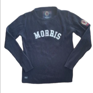 Marinblå ribbstickad tröja Morris - Ribbstickad marinblå tröja från Morris med vit broderad logga på bröstet och patch på ena ärmen. Långärmad och i klassisk, sportig stil. Perfekt för dig som gillar preppy vibes och snygga detaljer.