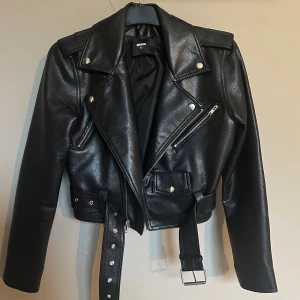 Svart skinnjacka från Bik Bok - Snygg svart skinnjacka från Bik Bok med klassisk bikerstil. Jackan har silvriga detaljer, dragkedjor, knappar och ett bälte i midjan. Långärmad och med cool krage, perfekt för att lyfta din outfit. Passar dig som gillar edgy och trendiga plagg. Ordinare pris 899kr. 