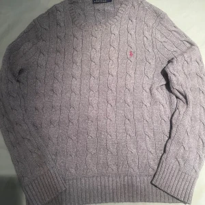 Ljusgrå kabelstickad tröja Polo Ralph Lauren - Kabelstickad tröja från Polo Ralph Lauren i ljusgrått med klassisk rund hals och broderad röd logga på bröstet. Tröjan är långärmad och har ribbade muddar och nederkant. Perfekt för lager-på-lager under höst och vinter.