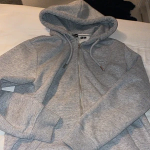 Grå hoodie från Superdry med dragkedja - Mysig grå hoodie från Superdry med broderad logga på bröstet. Tröjan har huva med snörning, hel dragkedja och mjukt fleecefoder på insidan. Perfekt för chill dagar eller när du vill ha något bekvämt och stilrent.