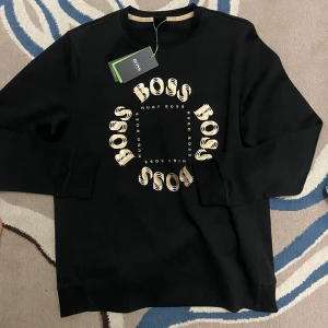 Svart sweatshirt från Hugo Boss - Svart sweatshirt från Hugo Boss Storlek M med rund hals och lång ärm. Framsidan har ett stort cirkulärt tryck med BOSS i guld och vitt, samt mindre Hugo Boss-texter. Tröjan är tillverkad i mjuk bomull och har ribbade muddar vid ärmslut och nederkant. Perfekt för en cool och avslappnad stil.