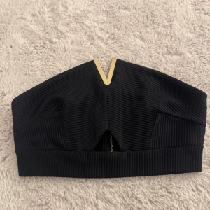 Svart bandeau tubtopp från Miss Selfridge - Säljer en svart bandeau tubtopp från Miss Selfridge med snygga ribbade detaljer och en guldig V-formad metall vid halsen.               Toppen har en guldfärgad dragkedja baktill och är axelbandslös.  NYSKICK - aldrig använd 