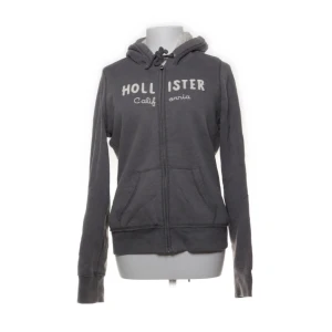 Grå hoodie från Hollister med päls - Mysig grå hoodie från Hollister med vit text på bröstet, huva med snörning och två stora fickor framtill. Tröjan har dragkedja och är tillverkad i bomull och polyester. Perfekt för chill dagar och snygg till jeans.