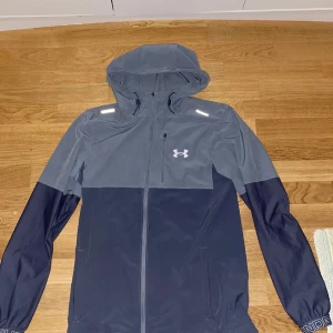 Blå och grå vindjacka Under Armour - Snygg vindjacka från Under Armour i två nyanser av blått och grått. Jackan har huva, hel dragkedja framtill, reflekterande detaljer på axlarna och logga på bröstet. Perfekt för träning utomhus när det blåser. Fickor med dragkedja och sportig look.