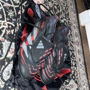 Adidas Predator fotbollsskor svart/röd - Säljer ett par Adidas Predator fotbollsskor i svart med röda och grå detaljer. Skorna har snörning, mönstrad ovandel och tydliga Predator-loggor undertill. Perfekta för dig som vill sticka ut på planen med snygg design och bra grepp. Bra skick men finns tecken på användning.