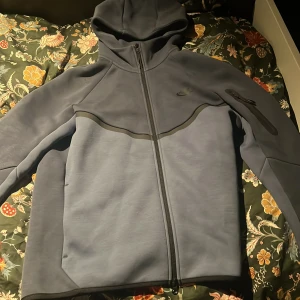 Nike Tech Fleece blå hoodiejacka M - Nike Tech Fleece hoodiejacka i mörkblå färg med hel dragkedja och huva. Jackan har en stilren design med svarta detaljer, Nike-logga på bröstet och en praktisk ficka med dragkedja på ärmen. Perfekt för dig som gillar sportig och clean stil.