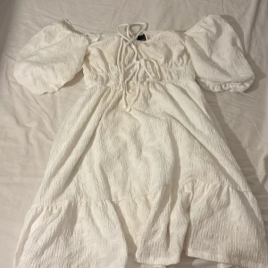 Vit kort klänning från Gina Tricot - Superfin vit kort klänning från Gina Tricot med puffärmar och smock i midjan. Klänningen har snörning framtill och volangdetaljer nertill. Perfekt för sommaren.💕