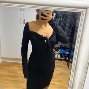 Svart omlottklänning med knyt - Snygg svart kortklänning med omlott och knyt framtill. Klänningen har långärmade ärmar och en v-ringad, offshoulder design som ger en trendig look. Materialet är mjukt och stretchigt, vilket gör att den sitter tight och framhäver figuren.