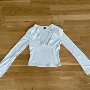 Vit långärmad topp med spets från SHEIN - Säljer en vit långärmad topp från SHEIN i storlek S. Toppen har en djup v-ringning med breda spetsdetaljer längs halsen och är figurnära i modellen. Materialet är mjukt och stretchigt, perfekt för dig som gillar romantiska vibes och snygga detaljer.