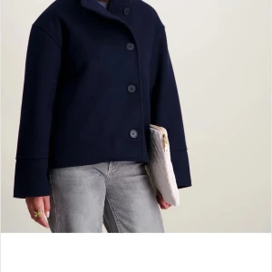 Mörkblå ulljacka från RYVLS Stockholm - Snygg mörkblå jacka från RYVLS Stockholm i ullmix. Jackan har rak passform, hög krage och stora knappar framtill. Perfekt för dig som gillar stilren och minimalistisk look. Passar till både jeans och kjol.