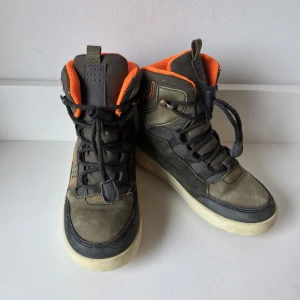 Grå höga sneakers från Ecco strl 30 - Snygga höga sneakers från Ecco i grått med orange detaljer och snörning. Skorna har en robust vit sula och är tillverkade i en kombination av skinn och syntet. Insidan är fodrad med orange tyg och de har praktiska snörlås för enkel påtagning.