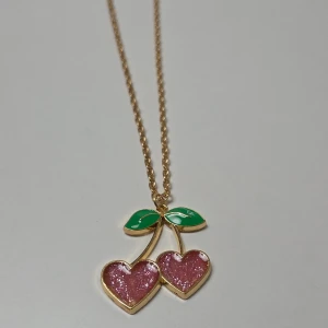 Halsband med glittrigt körsbärshänge, 45 cm - Gulligt halsband med ett hänge i form av två körsbärshjärtan i rosa glitter och gröna blad. Kedjan är guldfärgad och hänget har en söt och lekfull design som verkligen sticker ut. 45 cm + 5 cm förlängningskedja.