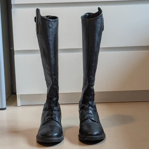 Ridstövlar i skinn från CRW - Säljer ett par klassiska svarta ridstövlar från CRW i äkta skinn. Stövlarna har snörning framtill, låg klack och rund tå. Perfekta för ridning och har en stilren design med diskreta detaljer och knäppning upptill. 2 små defekter på utsidan av ridstövlarna 