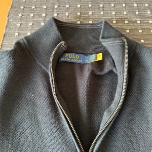 Svart half zip tröja Polo Ralph Lauren - Svart stickad tröja från Polo Ralph Lauren med half zip och klassisk ståkrage. Tröjan har lång ärm och den ikoniska broderade loggan på bröstet. Perfekt för dig som gillar stilrena och tidlösa plagg med snygg passform.
