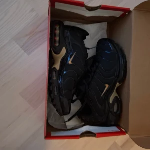 Nike tns svarta/guld sneakers - Nike Air Max Plus sneakers i svart med guldiga detaljer och synlig Air Max-dämpning i sulan. Skorna har en sportig vibe med vågformade linjer, mesh och syntetmaterial, samt snörning framtill. Swoosh-logga i guld på sidan och Tn Air-märke bak.
