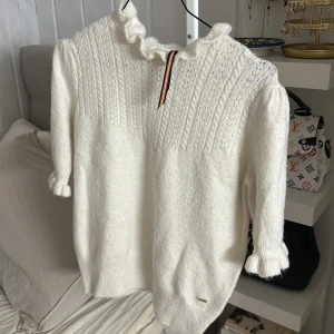Vit stickad tröja med volang Holly & Whyte - Superfin vit stickad tröja från Holly & Whyte i storlek S. Tröjan har snygga volangdetaljer vid hals och ärmslut, samt ett diskret mönster upptill. Mjuk och fluffig känsla, perfekt för kyliga dagar. Liten metallbricka med logga vid nederkant.