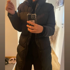 Svart lång pufferjacka med huva - Säljer en svart lång pufferjacka med huva och dragkedja framtill. Jackan har en i XS passform och är perfekt för kalla dagar. Den är quiltad och har långa ärmar för extra värme. Snygg och enkel att matcha med allt.