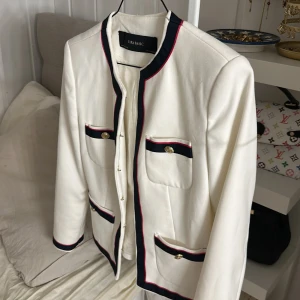 Vit kavaj med kontrastdetaljer Zara - Snygg vit kavaj från Zara Basic med marinblå och röda kontrastdetaljer vid kanter och fickor. Guldknappar på fickorna ger en lyxig touch. Rak passform, långärmad och tillverkad i ett mjukt, vävt material med sidenliknande foder. Perfekt för en chic och trendig look.