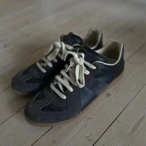 Maison Margiela gats sneakers svart - Maison Margiela sneakers i svart och mörkbrunt med beige detaljer. Skorna har ovandel i skinn och mocka, vita snören och klassisk gummisula. Ikonisk minimalistisk design med rund tå och lågt skaft. Perfekta för dig som gillar stilrena och tidlösa sneakers.