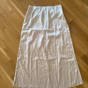 Vit/beige  långkjol - Säljer en stilren långkjol med rak passform. Elegant kjol i en ljus nyans. Kjolen har en slat och glansig yta, vilket ger den ett sofistikerat utseende. Passar perfekt för både vardag och fest. Aldrig använd behöver bara strykas.