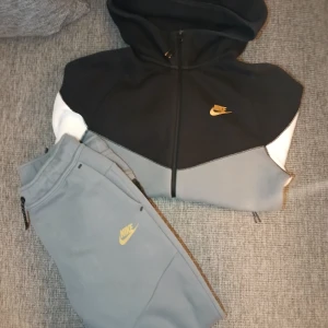 Nike hoodie och byxor svart/grå/vit - Nike hoodie  XS med dragkedja, svart upptill, grå på kroppen och vita ärmar. Gult Nike-logga på bröstet, två fickor med dragkedja och huva. Materialet är mjukt och känns som bomull/polyester. Perfekt för chill eller träning. Nike byxor L. Grå. Gult Nike-logga. Perfekt skick. Om ni vill se fler bilder skriv privat.