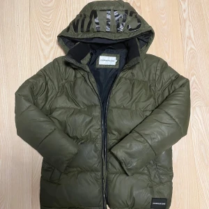 Grön pufferjacka från Calvin Klein Jeans - Säljer en grön pufferjacka från Calvin Klein Jeans med huva och stor logga på toppen. Jackan har dragkedja framtill, quiltad design och är helfodrad. Perfekt för kalla dagar och en clean look.