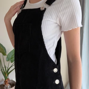 Svart manchesterklänning Stradivarius - Supersöt svart manchesterklänning från Stradivarius med klassiska hängslen, silverfärgade knappar och en stor ficka framtill. Kort modell med justerbara axelband och rak passform. Perfekt att styla med en t-shirt under för en chill look.