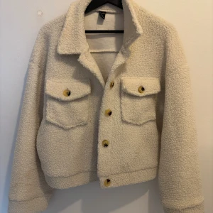 Off white  teddyjacka  - Mysig beige teddyjacka  med krage och två stora bröstfickor med knappar. Jackan har en croppad passform och stängs med guldfärgade knappar framtill. Perfekt för dig som gillar en chill och trendig look. Från Shein, passar XS om man vill ha lite oversized eller M där den sitter perfekt 
