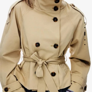 Beige trenchcoat med bälte och knappar - Beige kort trenchcoat från berskha storlek xs . Helt ny aldrig använd endast testad. Stilren beige trenchcoat med dubbelknäppning, höga axelpartier och bälte i midjan. Jackan har stora knappar, axelklaffar och justerbara ärmslut. Tillverkad i bomull och har en klassisk, rak passform som ger en snygg siluett.