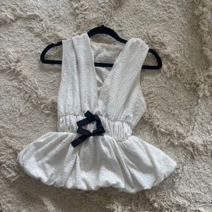 Vit omlott blus med rosett - Säljer en söt vit omlott blus med djup v-ringning och voluminös peplum nedtill. Blusen har en svart rosett i midjan som ger en extra detalj. Materialet är lätt och strukturerat, perfekt för vår och sommar. Ärmlös design för en fräsch look.