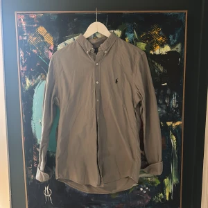 Grå skjorta från Ralph Lauren M - Snygg grå skjorta från Ralph Lauren i slim fit med klassisk button-down krage och broderad logga på bröstet. Skjortan är tillverkad i mjuk manchester och har långa ärmar med knappar vid manschetterna. Perfekt för dig som gillar stilrena och tidlösa plagg.